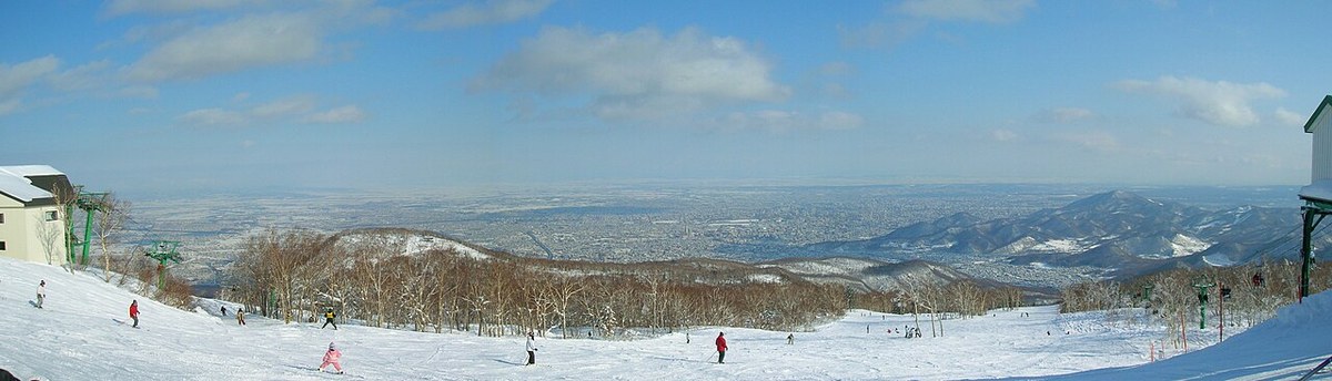 手稲山雪道上部的超寬全景,滑雪者散布在壓雪坡面上,左右各有一組吊椅纜車,遠方是整片札幌市區與延伸到地平線的平野