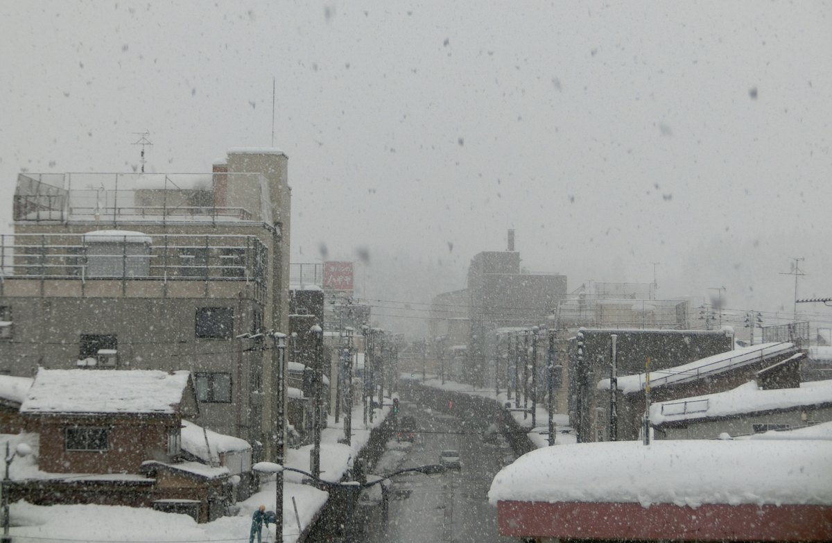 從高處俯瞰下雪中的十日町市區:兩排三四層的水泥樓房沿著一條筆直的街道往遠方縮小,每一戶的屋頂與雨庇上都壓著厚厚一層積雪,中間的車道被除雪成濕黑色、只有一兩台車,遠景整片被落雪糊成灰白。畫面左下角有一個穿藍色外套的人影,非常小