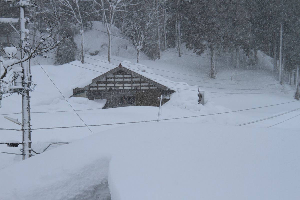 下雪中的山間聚落:一棟木造民家只剩三角形的屋頂與二樓的木格窗露在雪面上,一樓幾乎整個埋在雪裡;屋子前方是一整片平滑起伏的雪原,電線桿旁拉出的電線低到快貼著雪面。背景是積雪的緩坡與落葉喬木林,天空灰白,空中有明顯的落雪