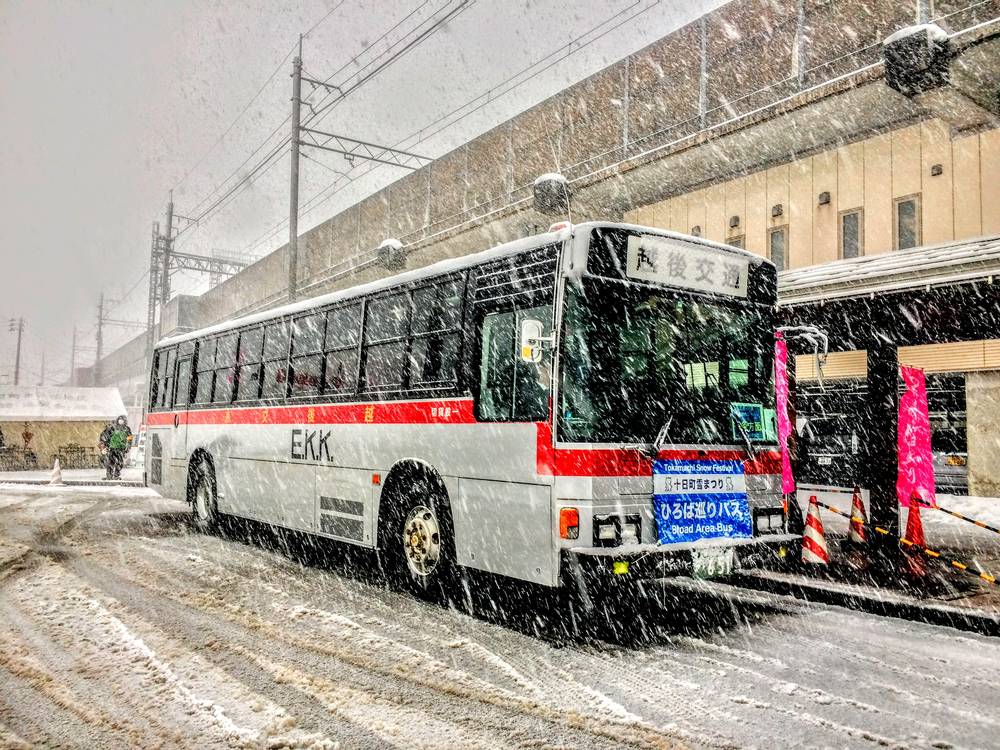 大雪中停在高架橋下的一輛白色越後交通路線巴士,車身側面漆著紅色橫線與「E.K.K.」字樣,車尾方向幕顯示「越後交通」;後保險桿上掛著一塊藍底白字看板,上排紅字寫「Tokamachi Snow Festival 十日町雪まつり」,下排大字寫「ひろば巡りバス Broad Area Bus」。巴士右側路邊立著三角錐和一面粉紅色的「ゆきまつり」直式布條,地面是被輾出輪痕的雪泥