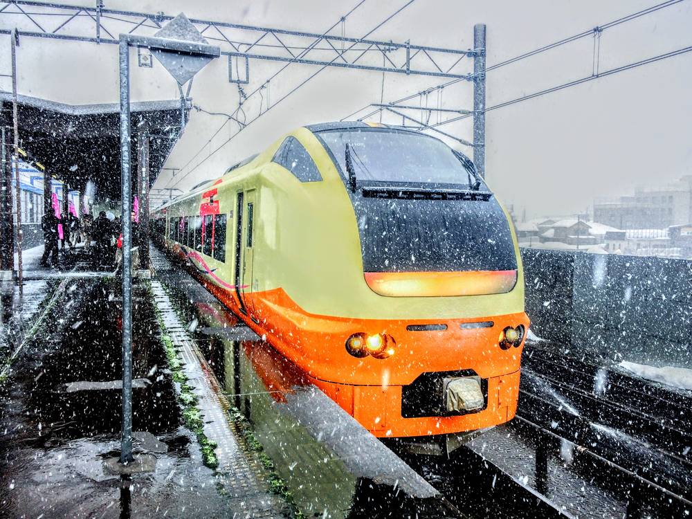 暴雪中停靠在十日町站月台的一列 JR E653 系電車,車頭是奶油色與橘色的流線造型,車頭燈亮著,車身側面也是橘色塗裝。月台雨庇邊緣與架空電線都掛著雪,左側月台遠處有幾個等車的人影與幾面粉紅色直式布條,畫面佈滿大顆雪花