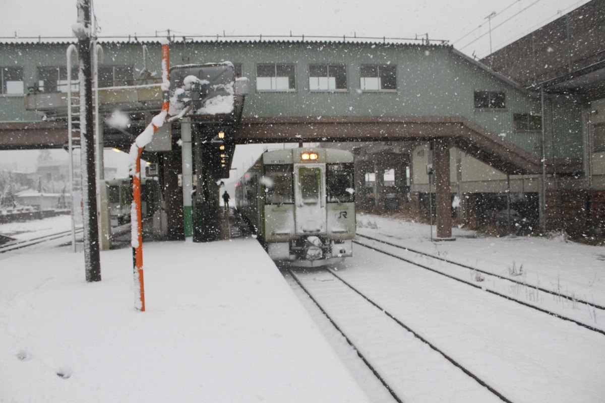 下雪天的十日町站月台:一列白綠相間、車頭掛著 JR 標誌的單節柴油車停在軌道上,前方軌道與月台之間積著未除的雪;月台上方跨著一座淺綠色鐵皮的跨線橋建物,橋下吊著亮著燈的看板。近景是一根積滿雪的橘白相間標柱,整個畫面佈滿飄落的雪花,遠處月台上只有一個模糊的人影