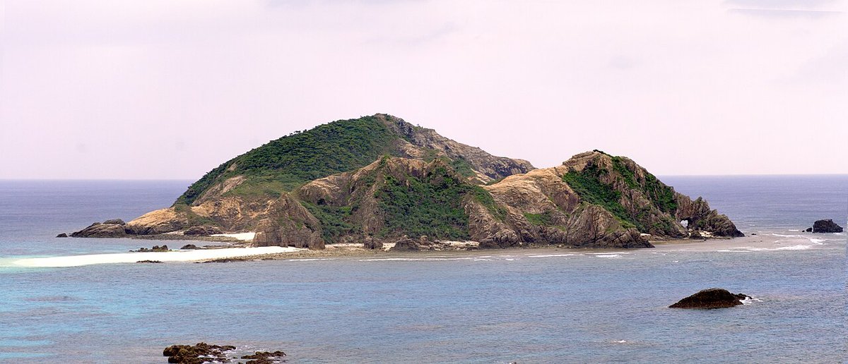 從渡嘉敷島阿波連海灘望出去的無人小島,島上一半綠樹一半裸露岩層,前方拉出一道白色沙洲與淺綠色礁岩淺灘,天空是一整片沒有藍色的灰白