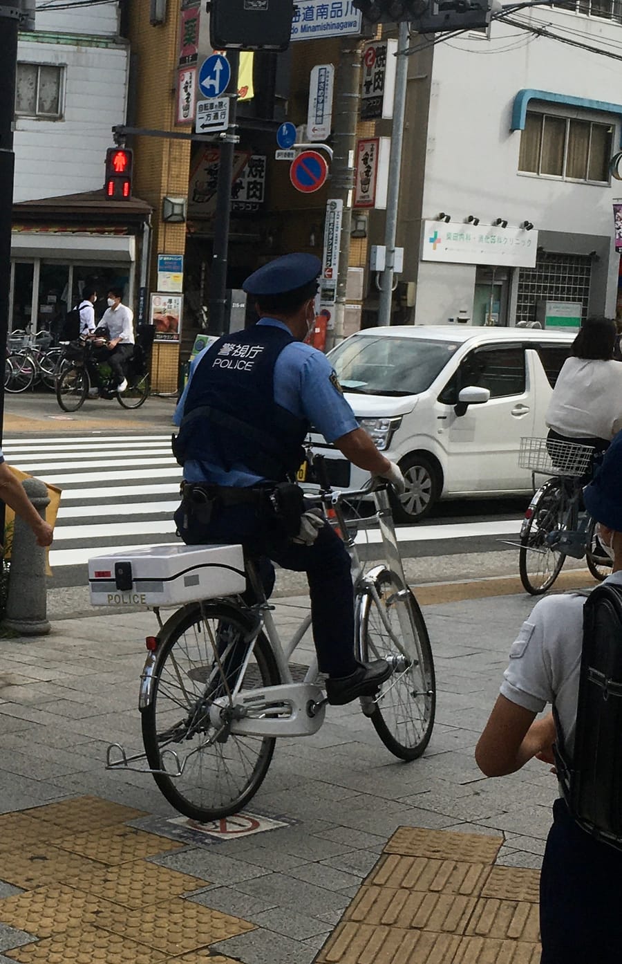 東京街頭路口:一名穿淺藍色制服、防彈背心背面印著「警視庁 POLICE」的警察跨坐在白色巡邏單車上等紅燈,車後架著一個印有 POLICE 的白色置物箱,斑馬線對面的行人號誌亮著紅色小人,旁邊另有一般民眾牽著單車
