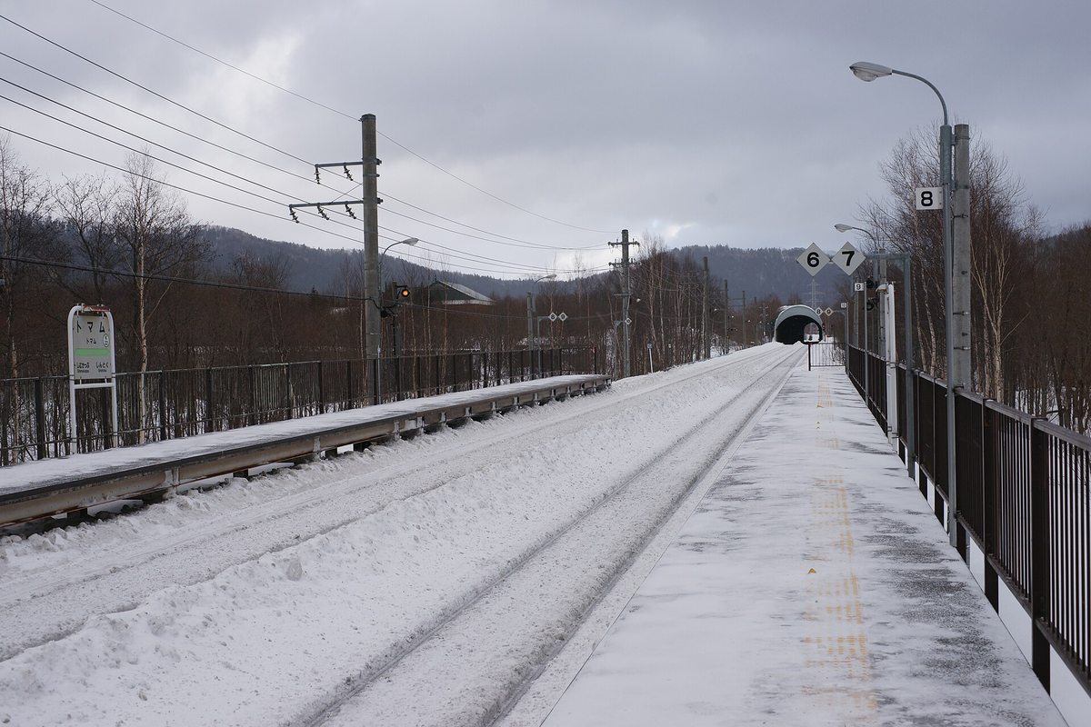 冬天的トマム駅 月台,路面積雪被踩實、鐵軌旁堆著除雪推起的雪堤,月台上空無一人,左側立著寫「トマム」的站名牌,前方軌道直直伸進一座隧道口,天空是厚重的灰雲