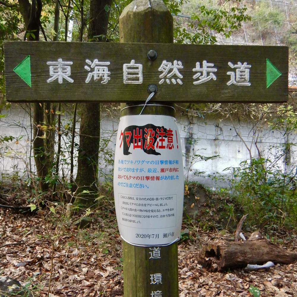 愛知縣瀨戶市東海自然步道的木製路標柱，橫木上刻著「東海自然歩道」與左右兩個綠色箭頭，柱身綁著一張護貝的「クマ出没注意！」告示，告示下緣印著 2020 年 7 月 瀨戶市