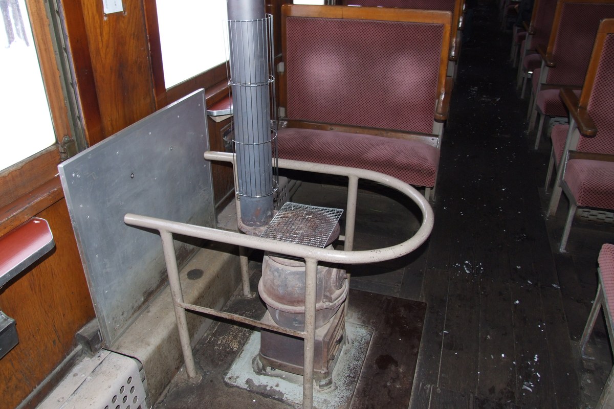 津軽鉄道暖爐列車車廂內部:走道旁立著燒煤的鑄鐵達磨火爐,外圍加了防護欄、上方接著煙囪,兩側是暗紅絨布對向座椅,窗外是雪原