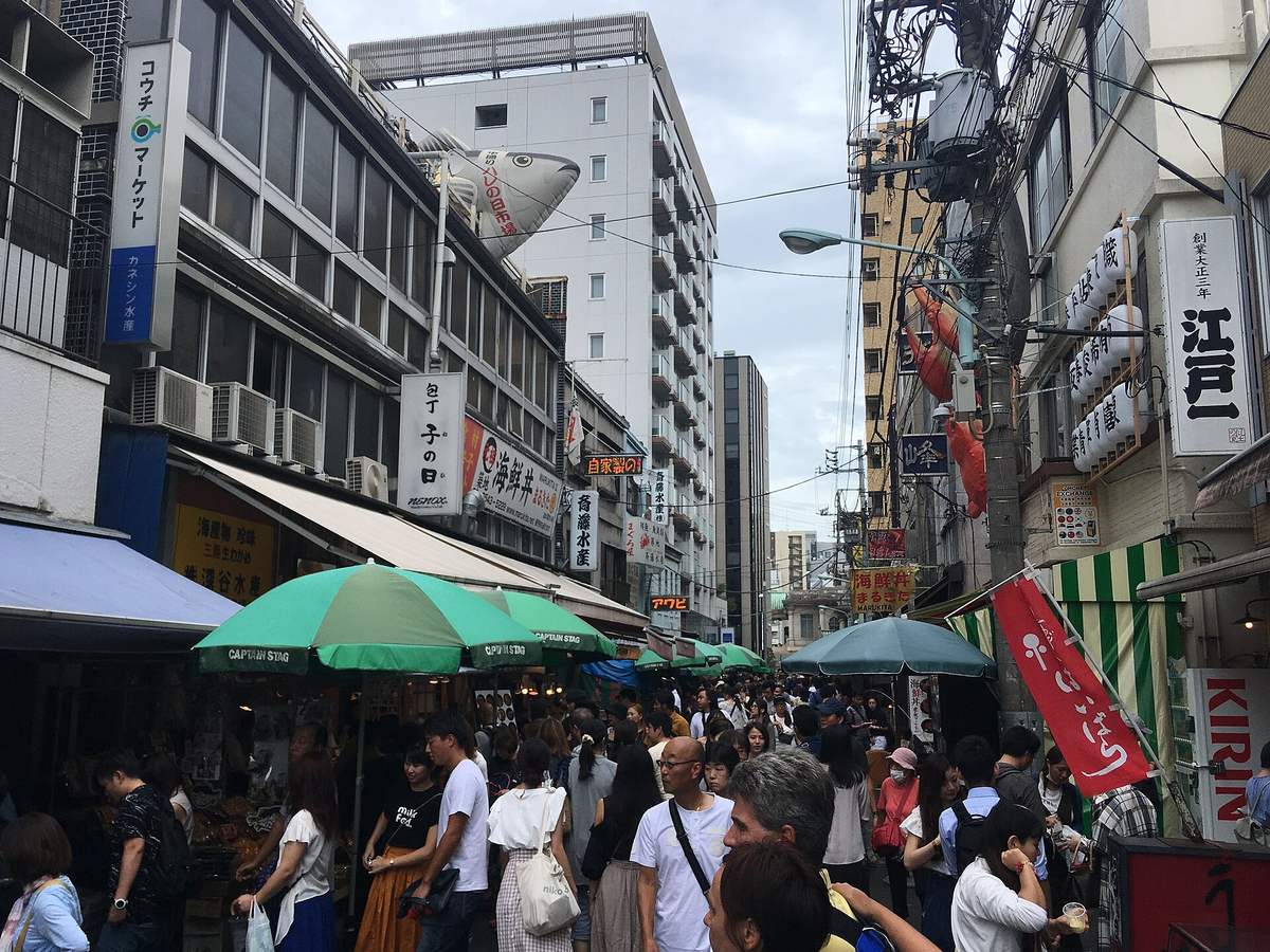 築地場外市場擠滿遊客的巷道,兩側是海鮮丼、水產與乾貨店舖,陽傘與遮陽棚一路排到街底