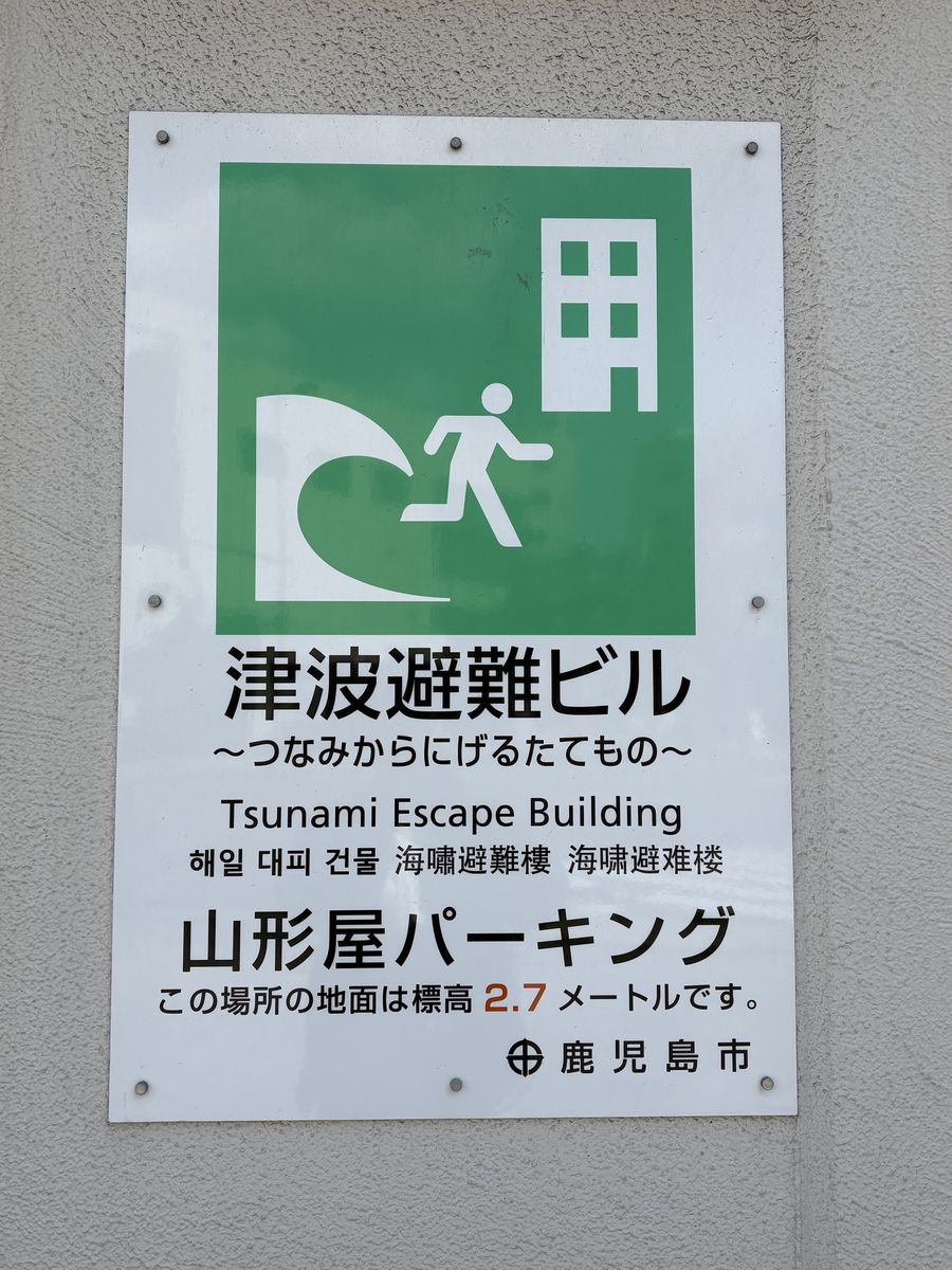 日本街頭牆面上的津波避難ビル標示牌：綠底白圖示畫著一道捲浪、一個奔跑的人形與一棟多層建築，下方依序以日文「津波避難ビル ～つなみからにげるたてもの～」、英文 Tsunami Escape Building、韓文、繁體中文「海嘯避難樓」、簡體中文標示，再往下是建物名稱「山形屋パーキング」與一行「この場所の地面は標高 2.7 メートルです。」，右下角印著鹿兒島市市徽