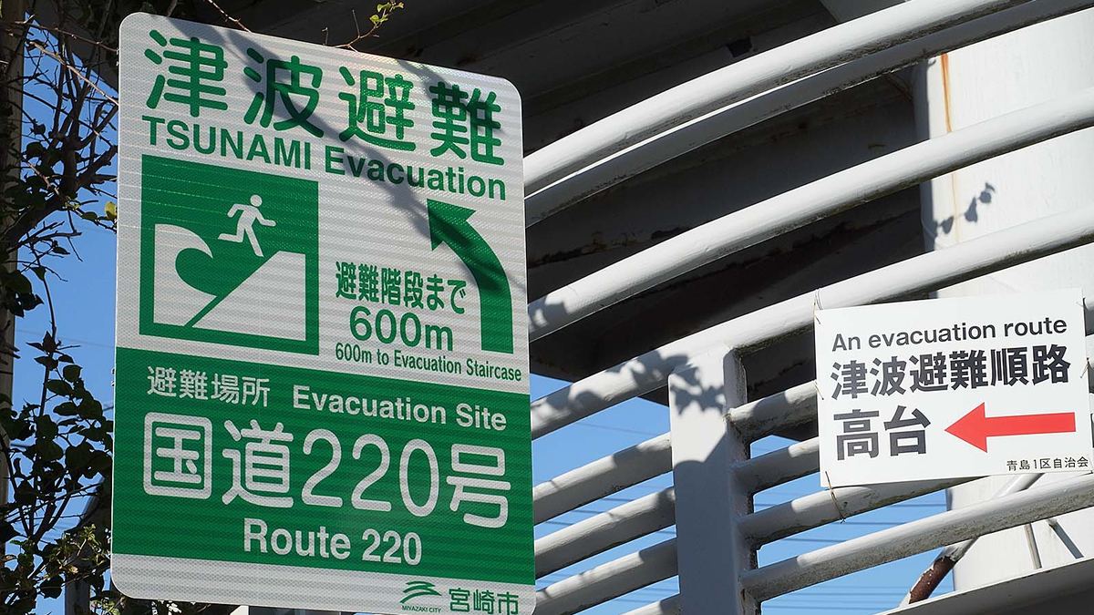 日本海岸邊的津波避難路標識，綠底白色波浪與奔跑人形圖案，箭頭指向高處避難方向