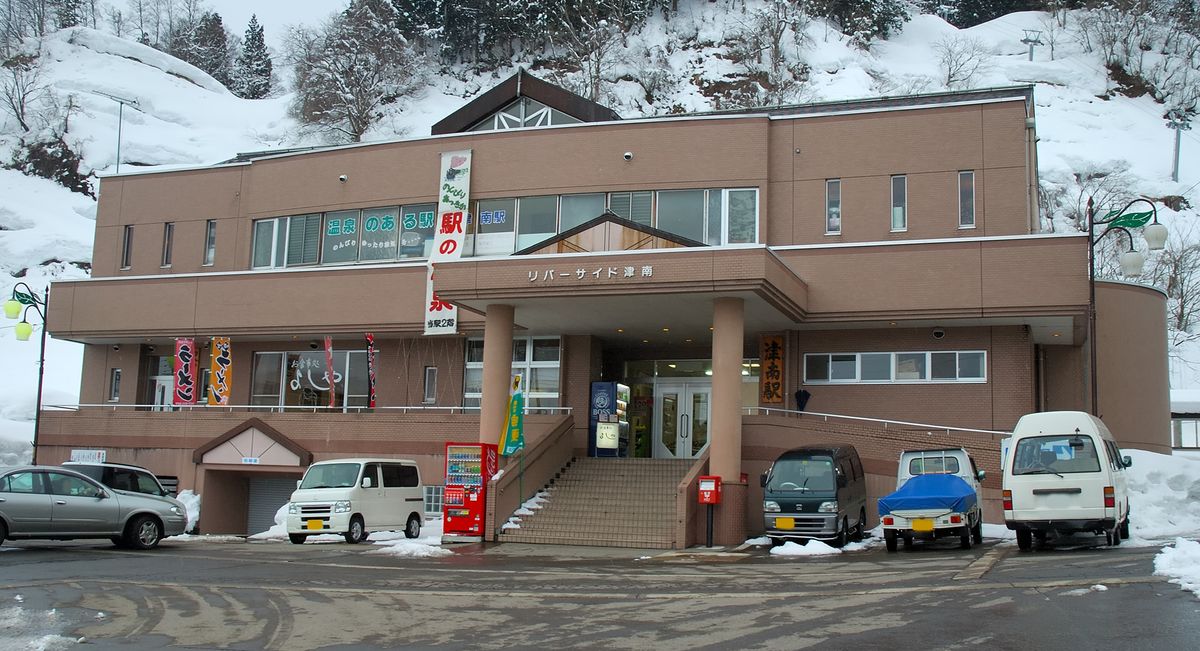 冬天的 JR 津南駅站房:一棟兩層樓的淺棕色建築,正中的門廊上寫著「リバーサイド津南」,入口右側掛著直式木牌「津南駅」,二樓窗上貼著「温泉のある駅」的字樣,門口旁垂著一條白色直式布條。入口是一段水泥階梯,旁邊有紅色自動販賣機與紅色郵筒。站前廣場的柏油路面是濕的,路邊殘留剷過的雪,停著幾輛廂型車與輕型貨車;建物背後是一整片積雪的山坡與樹林,天空灰白