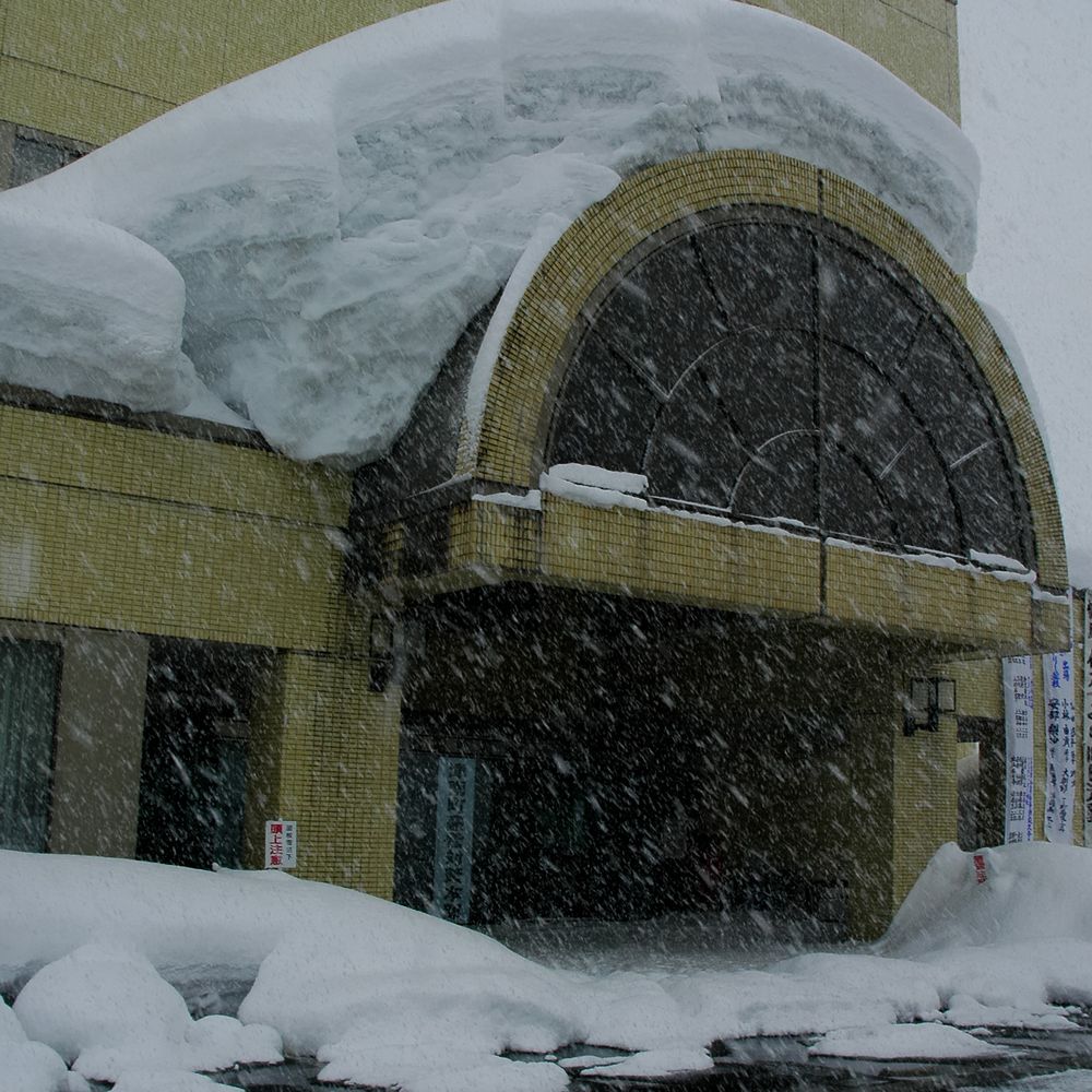 大雪紛飛中的津南町役場正門:米黃色磁磚外牆的建物,入口上方是一座半圓形拱窗,拱頂與旁邊屋簷上堆著厚厚一層積雪,積雪邊緣像一整塊白色厚板懸在屋簷外;入口左側牆上貼著一張紅字的「頭上注意」小告示,右側掛著幾條直式布條。前景是剷到路邊的雪堆與濕黑的地面,整個畫面佈滿斜斜落下的雪花