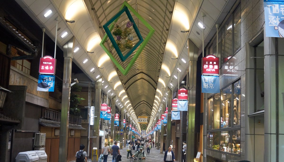 京都寺町京極商店街:拱形的鋼骨玻璃屋頂一路延伸到街底,兩側掛著紅色與藍色的店家旗幟與店招,街上零星幾個人走著,完全淋不到雨
