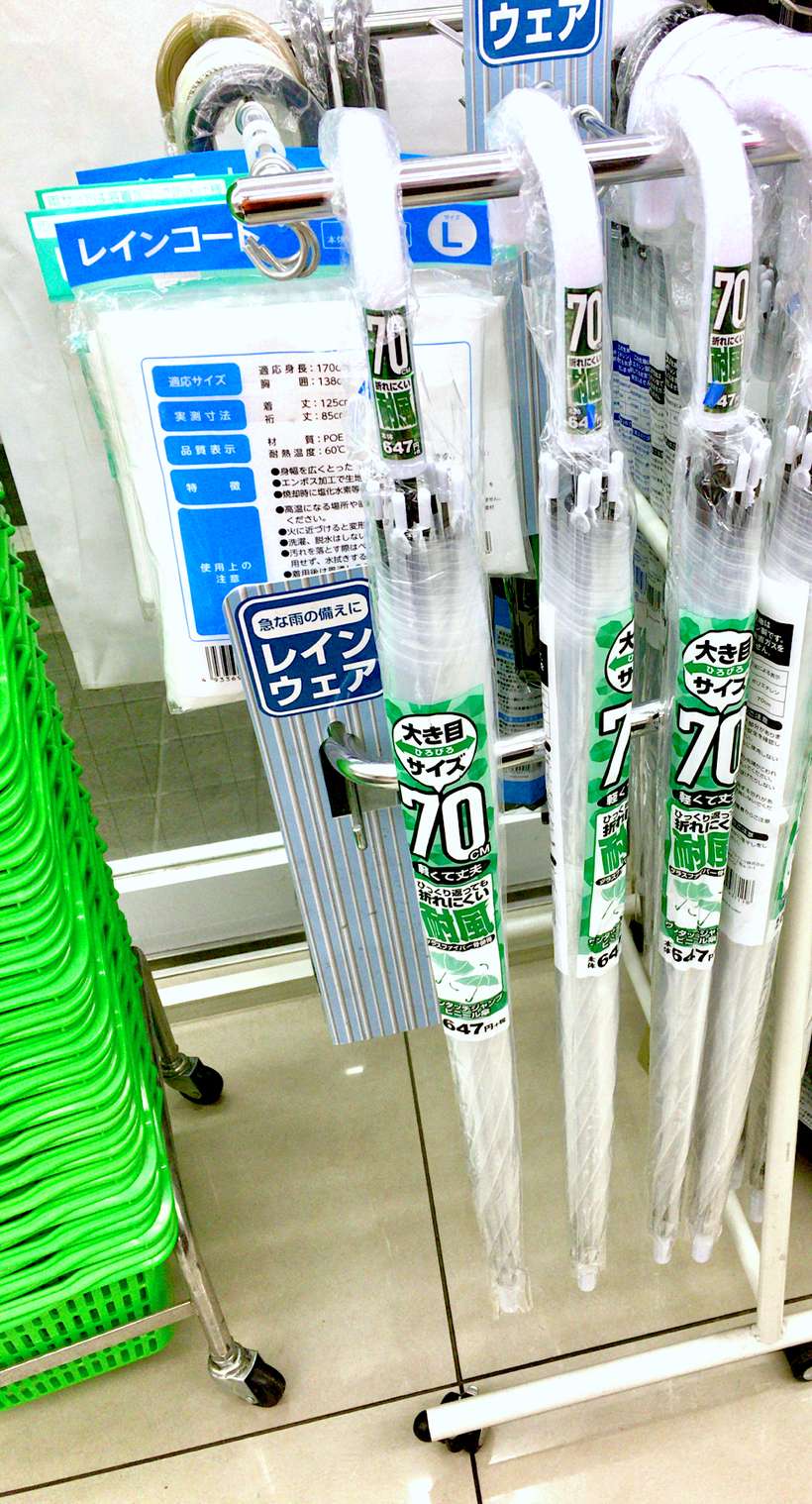 日本超商店內的傘架:三把包著塑膠套的透明傘並排掛著,套上印著「大き目サイズ 70cm」與「耐風」,後方貨架掛著成人用雨衣的包裝袋與藍色的レインウェア標示