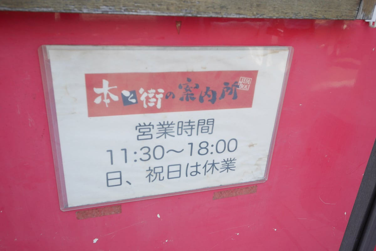 日本東京神保町一家店門口的粉紅色牆面上掛著塑膠框營業時間牌,牌子上半是紅底白字的「本と街の案内所」店名,下半印著「営業時間 11:30〜18:00」與「日、祝日は休業」