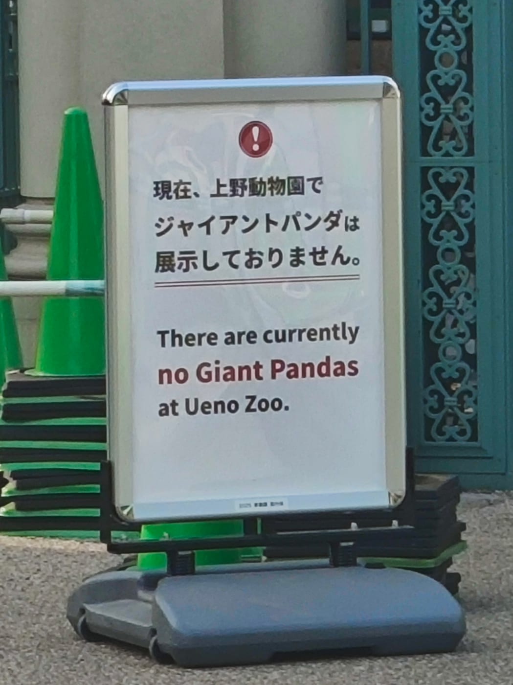 上野動物園門口的銀框 A 字立牌,白底黑字上方有紅色驚嘆號圖示,日文寫「現在、上野動物園でジャイアントパンダは展示しておりません。」,下方英文寫 There are currently no Giant Pandas at Ueno Zoo,牌子右側是青綠色鑄鐵門扇,左後方堆著綠色三角錐