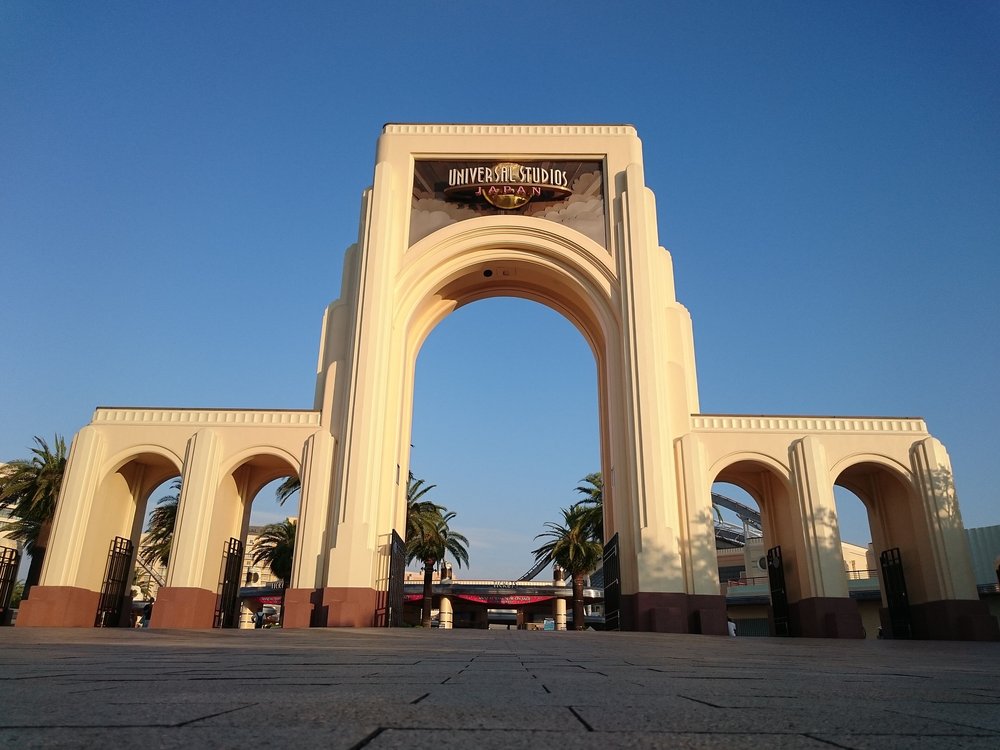 USJ 入口的米白色大拱門,頂端招牌寫 UNIVERSAL STUDIOS JAPAN,兩側小拱門下是黑色鐵柵門,傍晚藍天下廣場空無一人