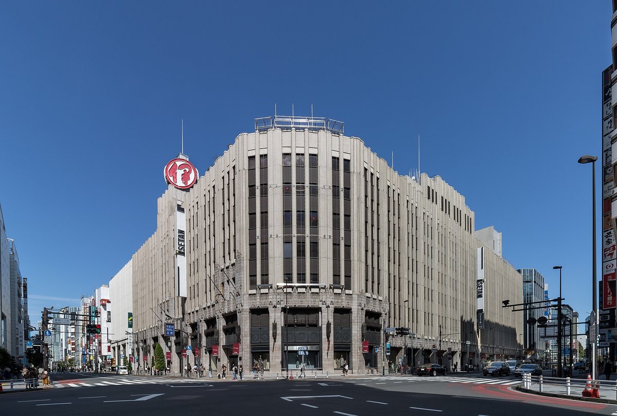 伊勢丹新宿店本館外觀:米色石造的裝飾藝術風建築佔滿新宿三丁目路口轉角,屋頂立著紅色圓形伊勢丹標誌,外牆垂著白底黑字 ISETAN 直幅,騎樓下是深色金屬門面