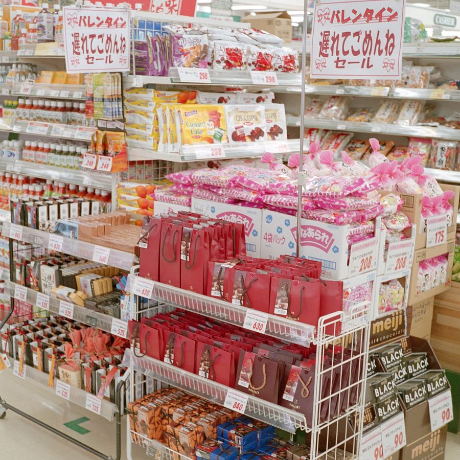 日本超市的情人節花車:兩塊白底紅字看板寫著「バレンタイン 遅れてごめんねセール」,鐵推車上層排著紅色與酒紅色的巧克力提袋、標價 630 與 840 円,右側紙箱裝著 meiji BLACK 與牛奶巧克力,標價 90 円