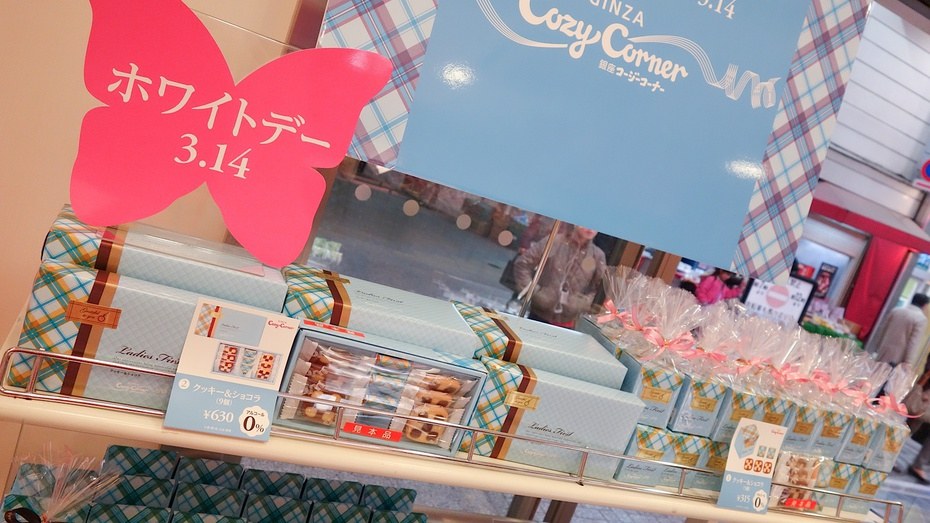 洋菓子店的回禮賣場:粉紅色蝴蝶造型立牌印著「ホワイトデー 3.14」,下方銀色鐵架上排著淺藍格紋包裝的長方形禮盒,中間一盒打開展示裡面的餅乾,旁邊價卡寫著クッキー&ショコラ(9個)¥630