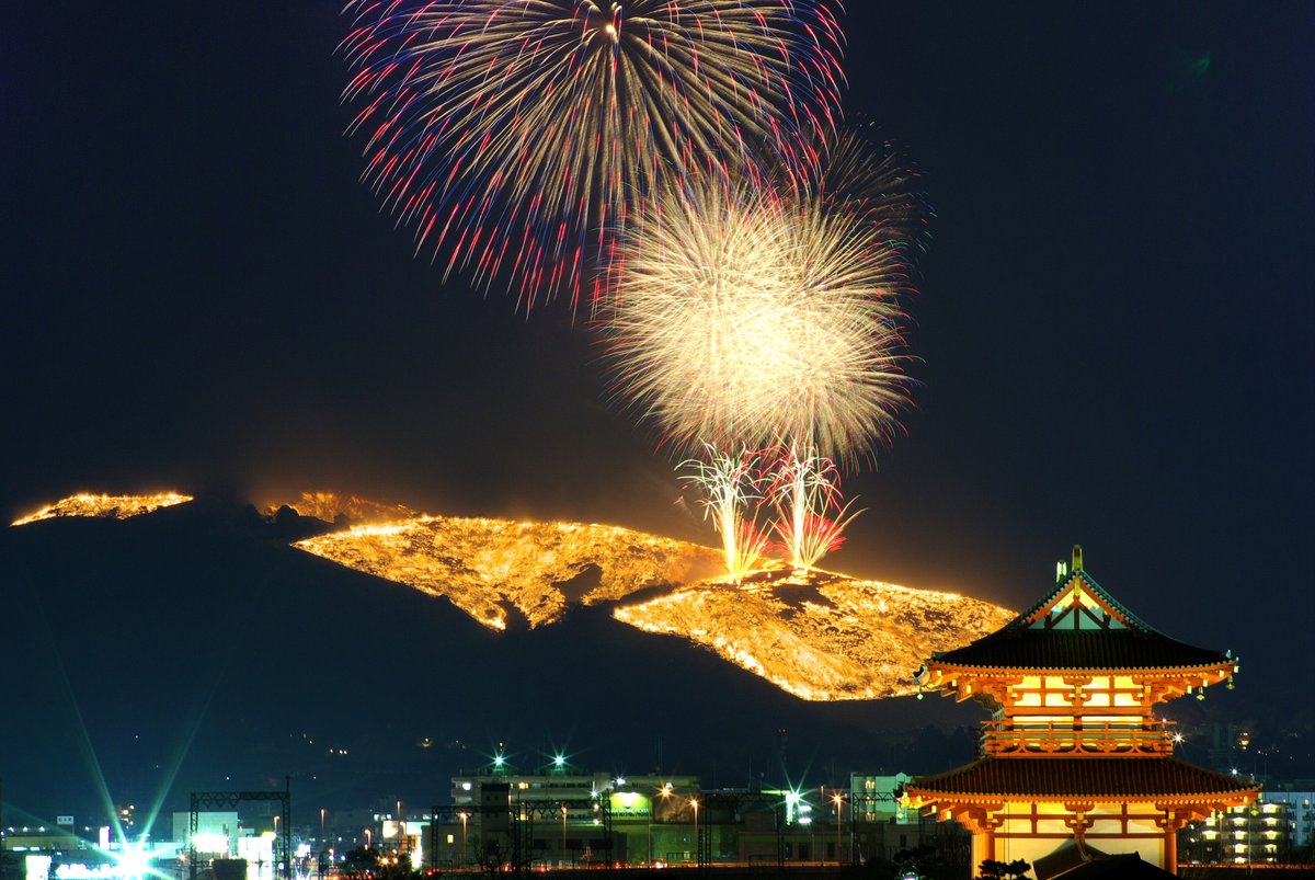 長時間曝光拍下的若草山燒山夜景:山坡上的火線連成一整片金黃色火面,上方同時炸開兩顆大花火,右下角是亮燈的平城宮跡朱雀門