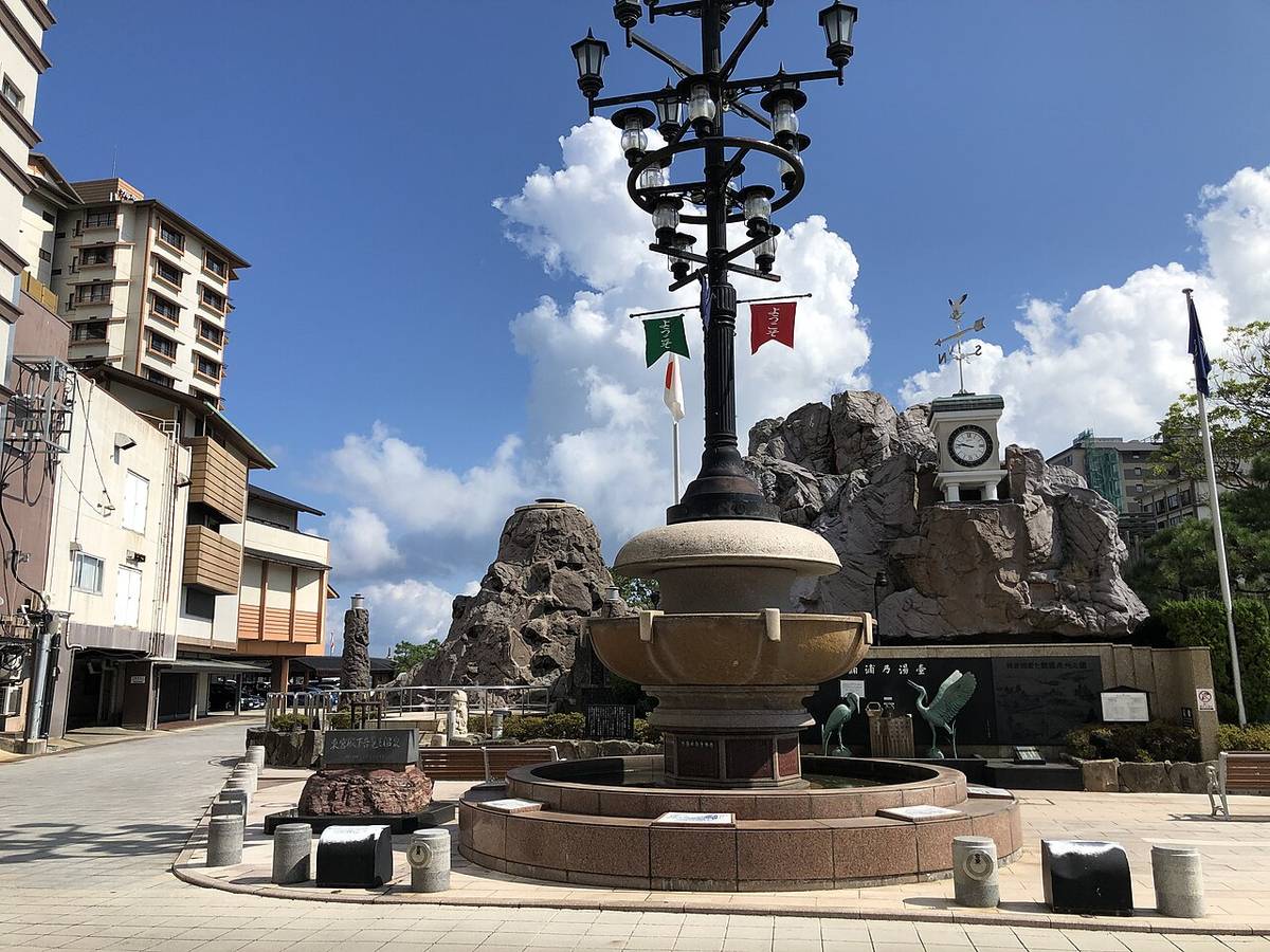 和倉溫泉街上的公共廣場,中央是一座圓形石砌水池與一根掛著多盞燈的鑄鐵路燈柱,柱上垂著兩面寫著「ようこそ」的小旗,水池後方是人造岩景與一座白色小鐘塔,兩旁是溫泉街的房子,地面鋪磚,沒有行人