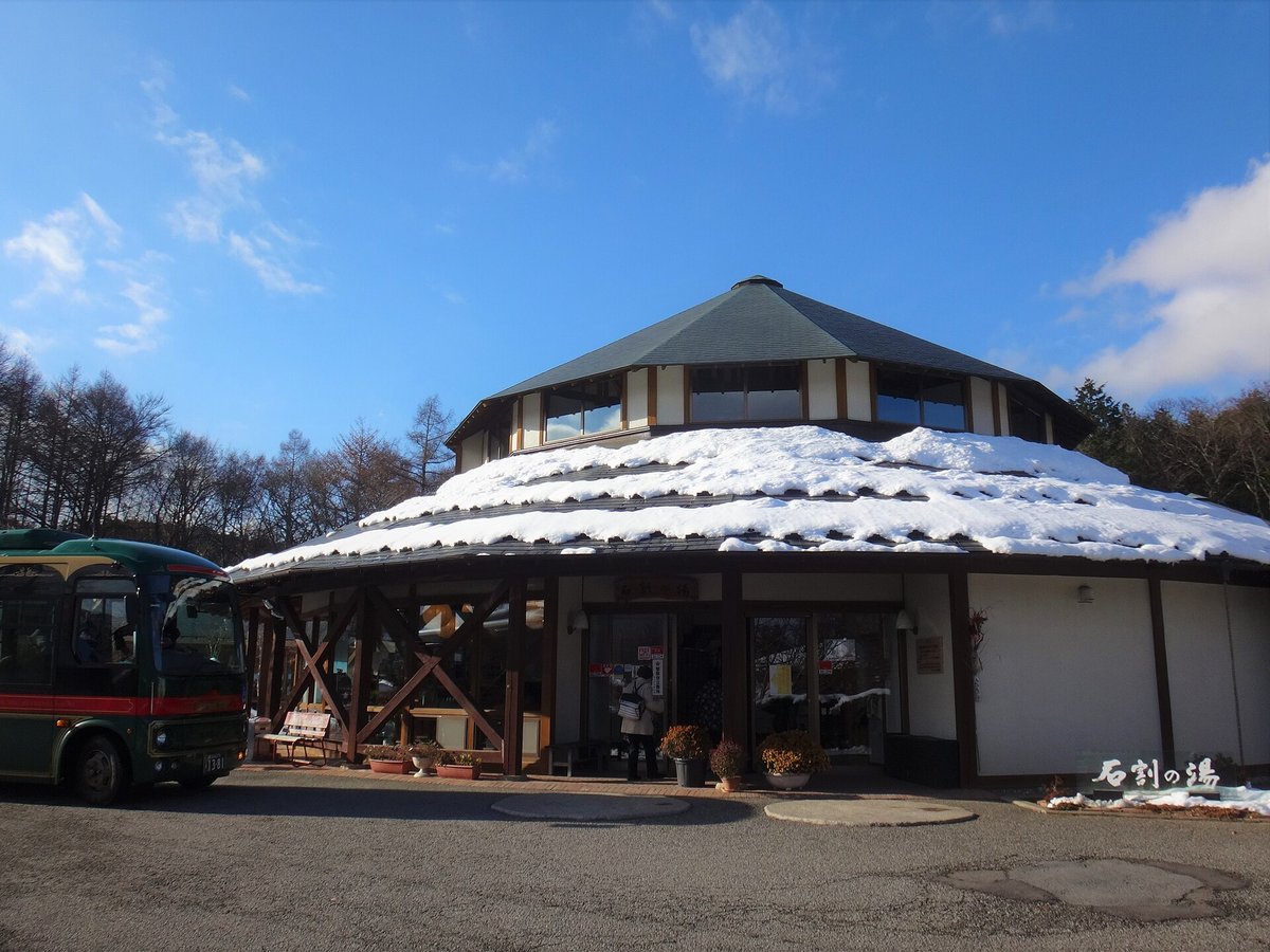 石割の湯的正面:八角形屋頂的溫泉建築,下層屋簷蓋著一圈積雪,右下角有「石割の湯」字樣,一位客人背對鏡頭站在入口,左邊停著一台墨綠色巴士