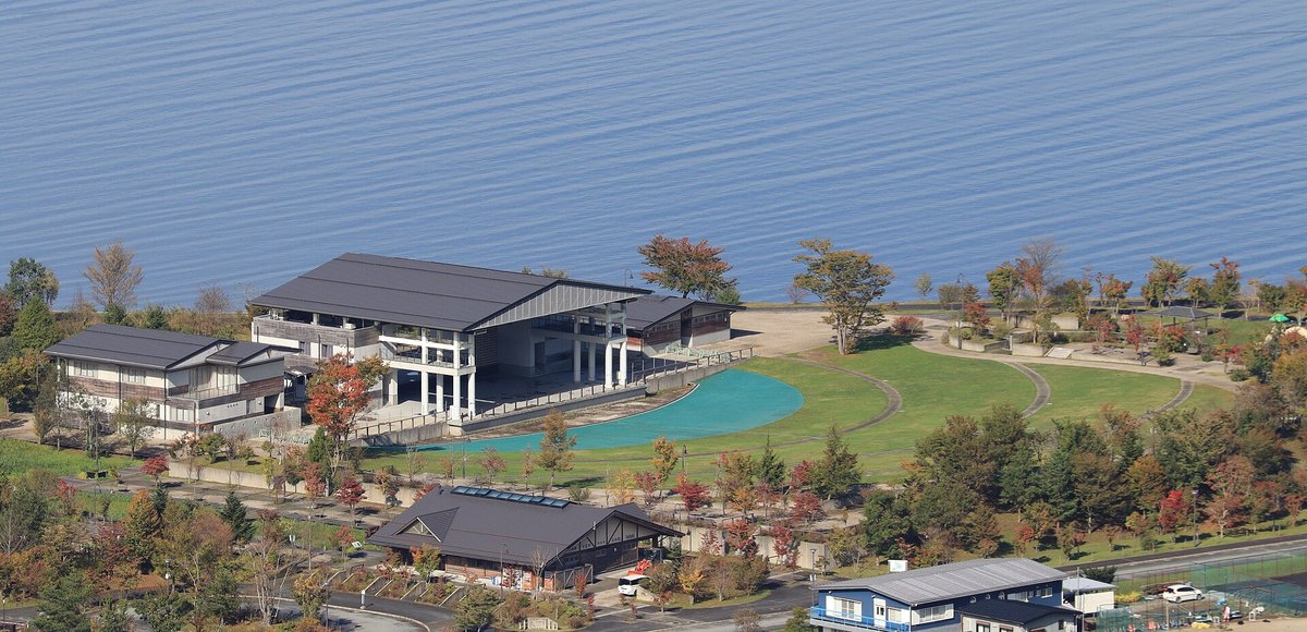 從山上俯瞰山中湖交流プラザきらら:湖邊幾棟深灰屋頂的建築、一片藍綠色的淺水池和弧形草坪步道,草坪外緣是淺色沙灘,上半部整片是湖面
