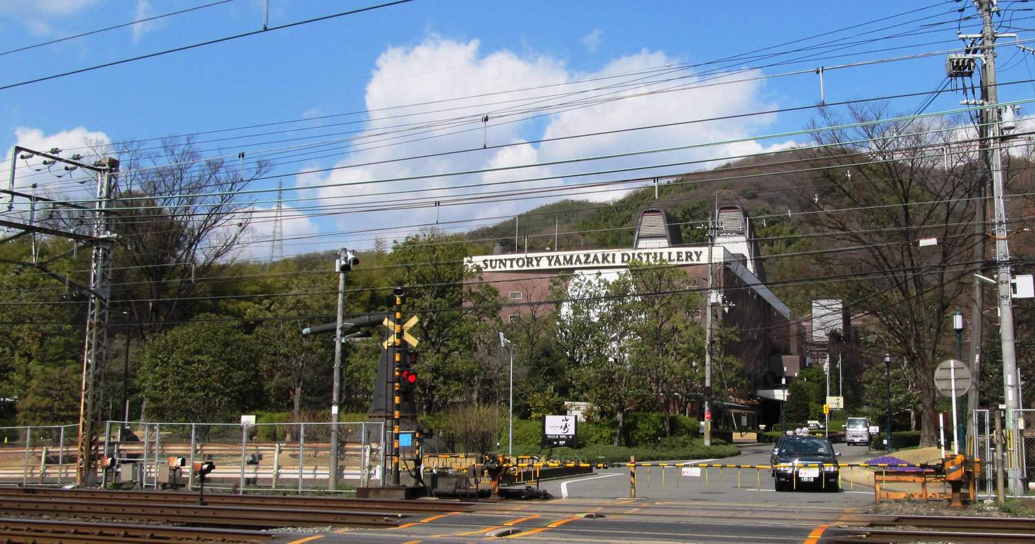 蒸溜所門口的鐵道平交道:畫面下緣是兩組鐵軌與塗成橘黑相間的道口鋪面,中央立著黃黑條紋的平交道警報機與交叉標誌,紅燈亮著、遮斷桿放下;桿子後面一輛黑色計程車與一輛白色小貨車停在通往蒸溜所的路上等待,那段路沒有人行道,只有一條白色路邊線;再往後是寫著 SUNTORY YAMAZAKI DISTILLERY 的紅褐色廠房與長滿樹的山坡,天空有電車架線與電線橫過