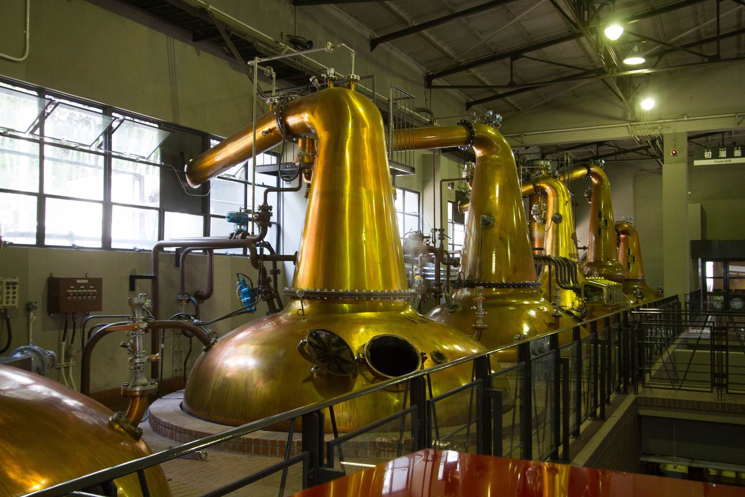 蒸溜棟二樓的走道視角:四座銅製蒸餾釜沿著磚砌基座一字排開,每座都是渾圓的鍋身接一段細長頸子,頂端彎出一根粗銅管往牆上接去;釜身被天花板燈打得發亮,左邊是一整排鐵框長窗,右方牆上掛著一塊寫著「初溜釜 Wash Still」的藍底標示牌,走道邊架著黑色欄杆,畫面裡沒有人