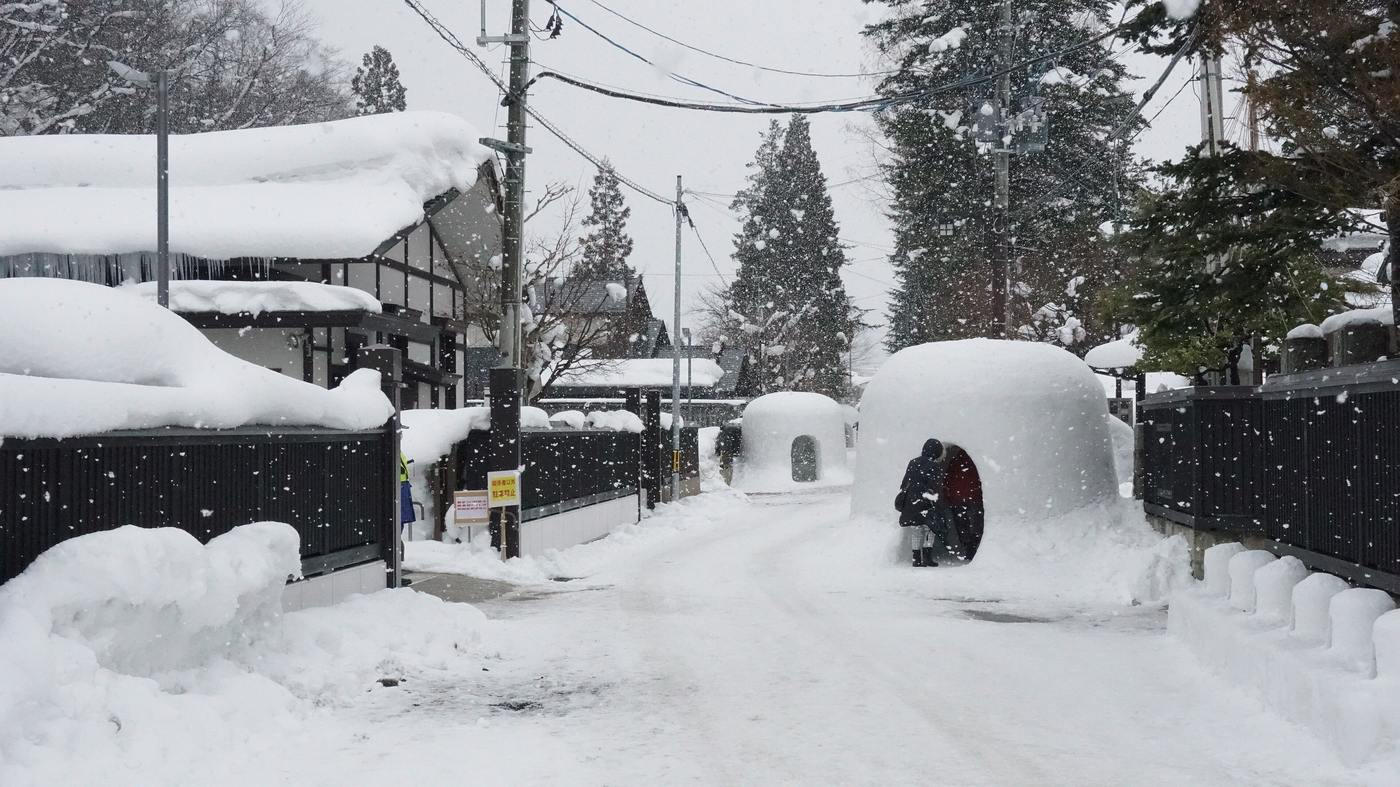 大雪紛飛的橫手羽黑町武家屋敷通，鏟過的窄路兩側是黑色板牆與積雪的屋頂，路旁立著兩座雪屋，一名遊客蹲在其中一座的入口