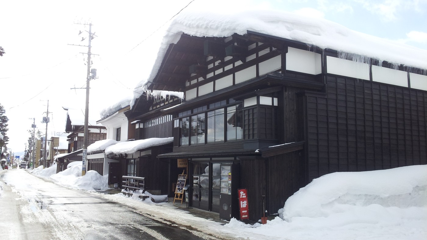 橫手中七日町通的冬景：一棟黑木造町家屋頂頂著厚厚一層雪、屋簷垂下長冰柱，路面已鏟成濕黑色，兩旁的雪堆高到店家門面一半