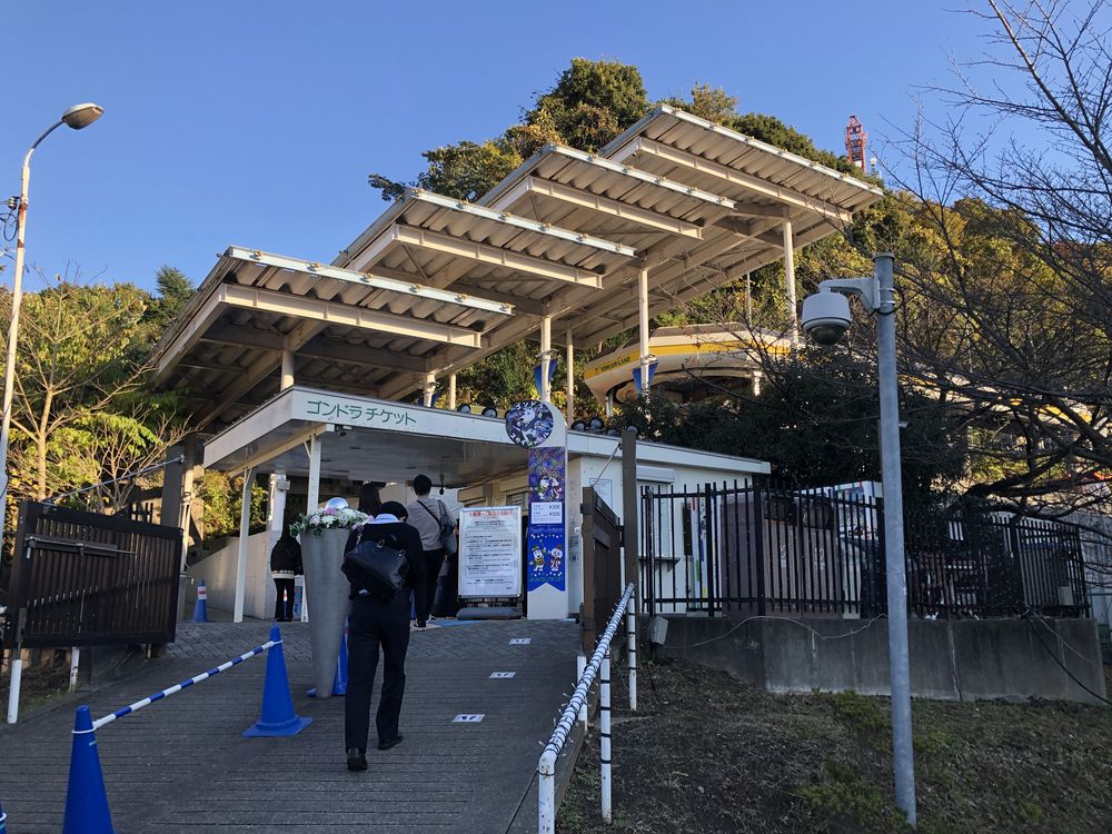 纜車京王口站的售票處入口:屋簷上寫著ゴンドラチケット,乘客背對鏡頭沿坡道排隊走進去,右邊直立看板印著單程與來回票價