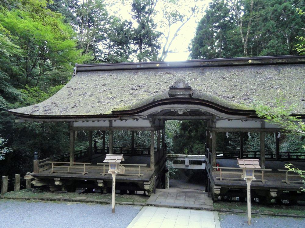 由岐神社的拜殿:長滿青苔的大屋頂,建築從中間分成左右兩半、留一條石板通道穿過去,兩側是四面敞開的木造高床,前方立著兩根小燈籠造型的木標柱,四周是杉林