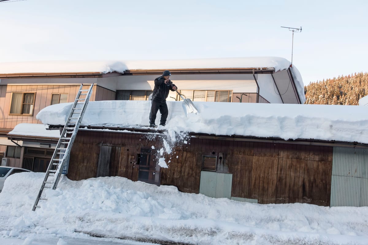 雪國住宅區的雪下ろし作業:一名居民架鋁梯爬上屋頂,用雪鏟把厚達一公尺的積雪推落到路旁