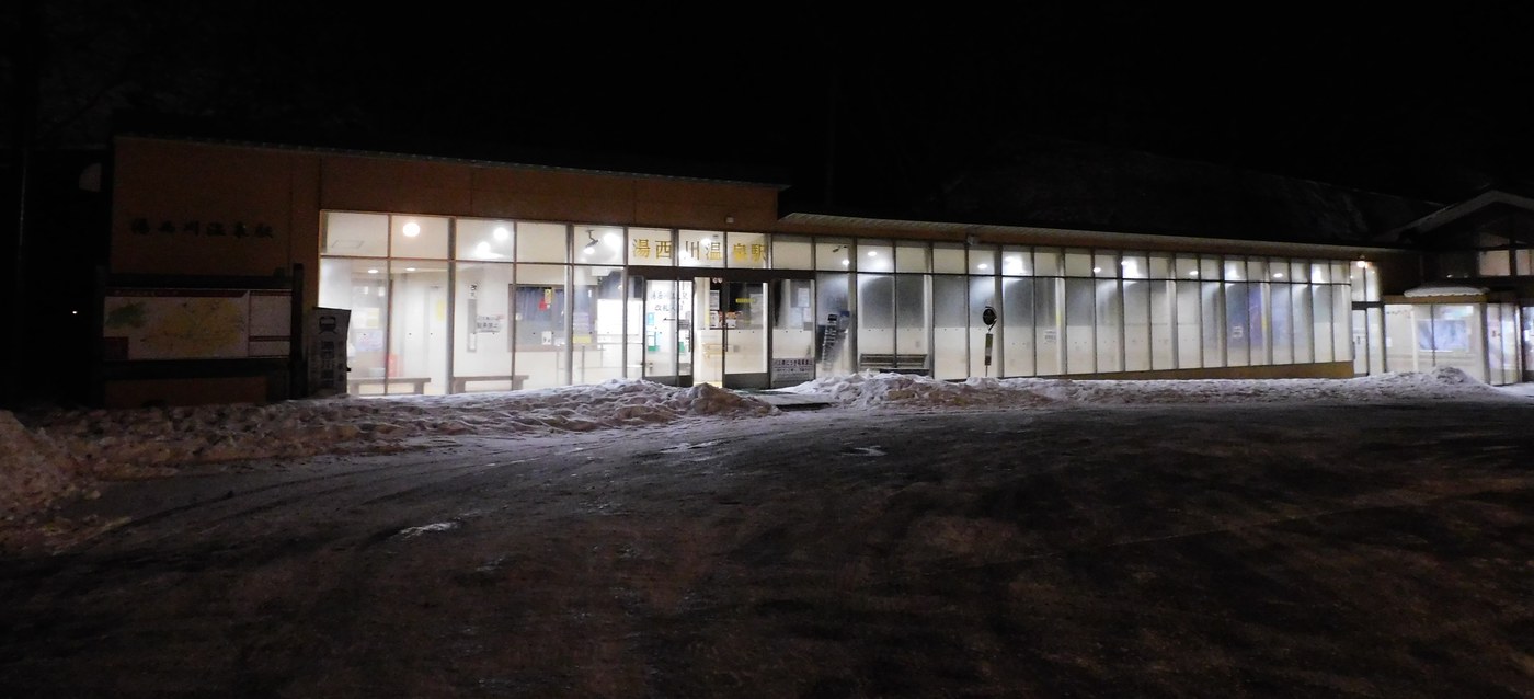 冬夜的湯西川温泉駅站房,玻璃外牆亮著燈,站前的雪剷到兩旁,門口右側立著巴士站牌