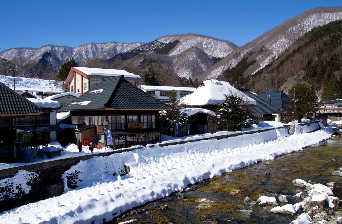晴天雪後的湯西川溫泉街,清澈的河流與堆滿積雪的河岸,對岸是黑瓦與覆雪茅草屋頂的木造房子,背後是積雪的山
