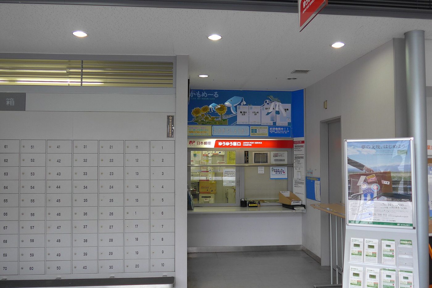 日本郵局內的小型窗口,紅底白字招牌寫著「ゆうゆう窓口 JAPAN POST SERVICE COUNTER」與 日本郵便 標誌,窗口內側檯面堆著數個牛皮紙箱與白色包裹,牆上貼著告示,左半邊是整面編號 1 到 70 的私書箱信箱,右側立著ゆうちょ銀行的直立式看板
