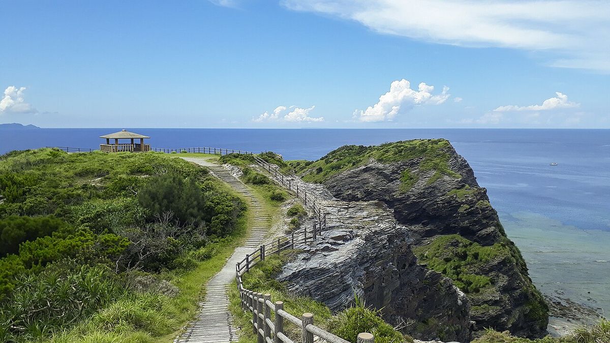 座間味島女瀬の崎展望台,草坡頂端一座木造六角涼亭,一條有木欄杆的步道沿著岩石海岬往上走,右側是斷崖與整片深藍海面,遠方水平線上浮著幾座島影