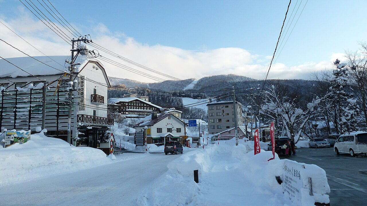 蔵王温泉冬季街道，路面壓雪、兩側堆起及腰雪堤，木造旅館與民宿沿街而立，上方橫過纜車纜線，遠方是覆雪山坡