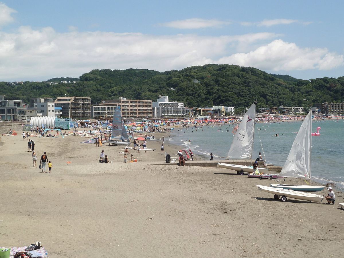 夏季的逗子海岸:前景是空曠的褐色沙灘,中段沿著海岸線排開一整列海之家的帳篷與彩色遮陽傘、遠處有零星戲水的人影,右側沙上停著三艘帆船,海灣後方是低矮的綠色丘陵與白色公寓大樓